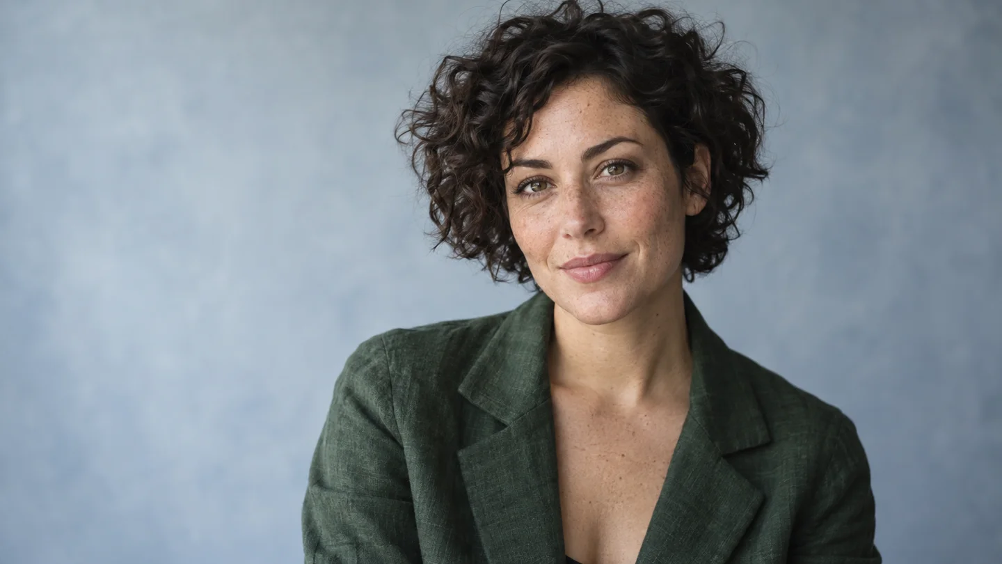 A woman with short dark curls and a green jacket against a pale studio background.